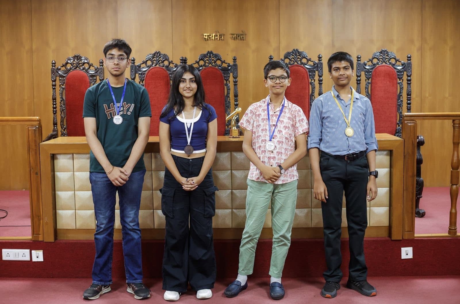 Four students with medals standing in front of tall red chairs